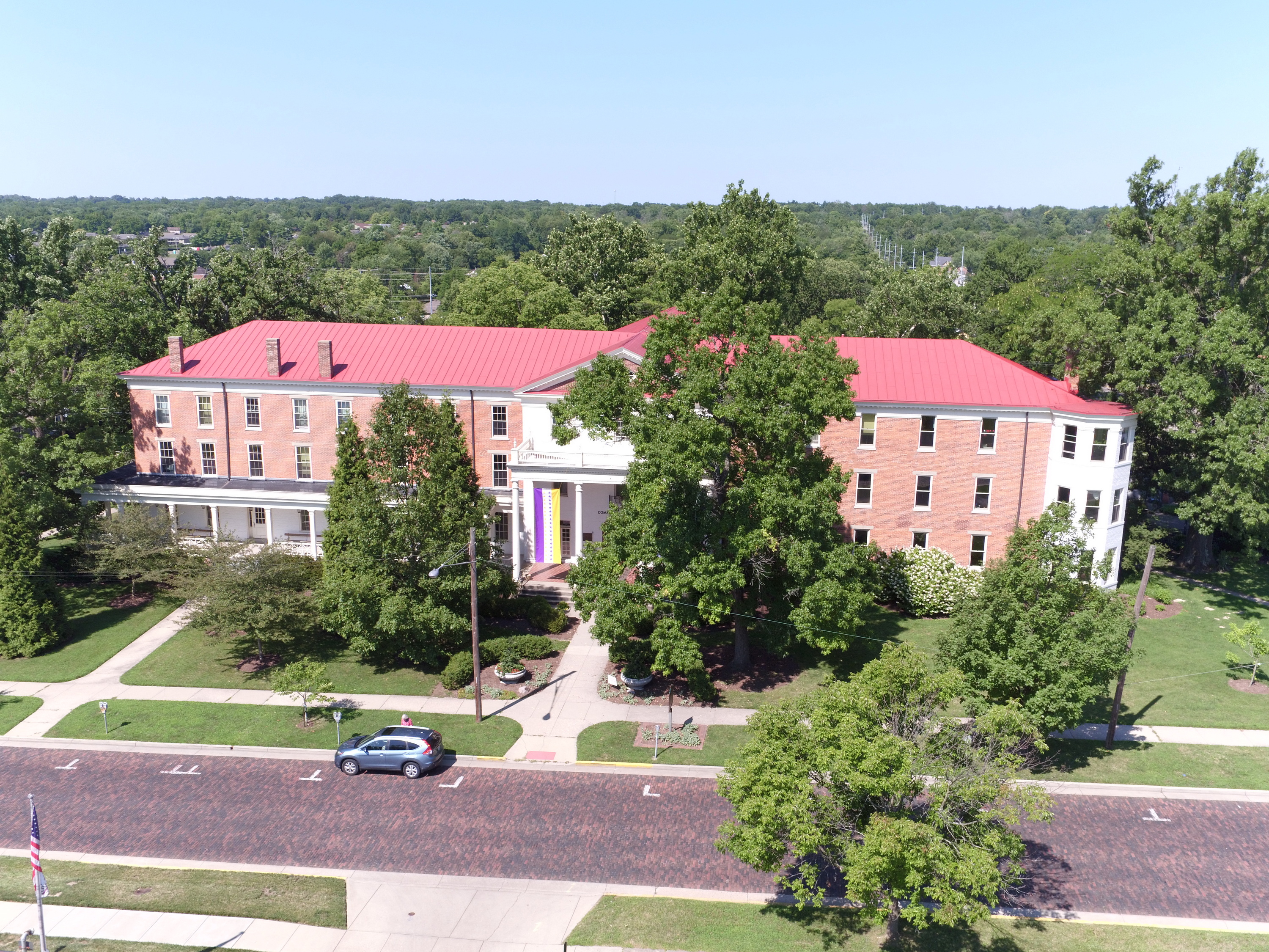 Red-roofed brick building surrounded by trees and a road.