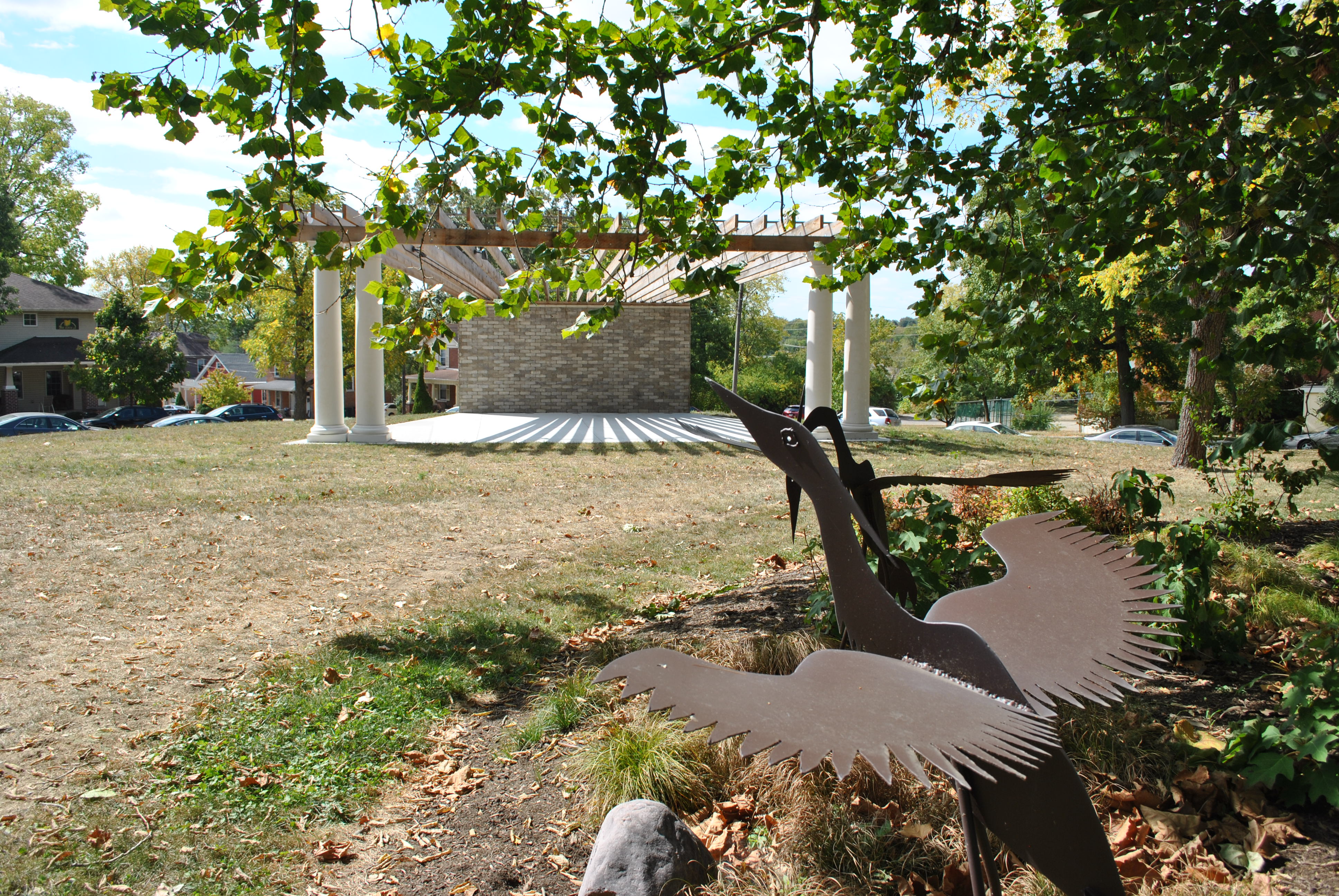 Sculpture of metal birds in a sunny park with trees and a pavilion in the background.