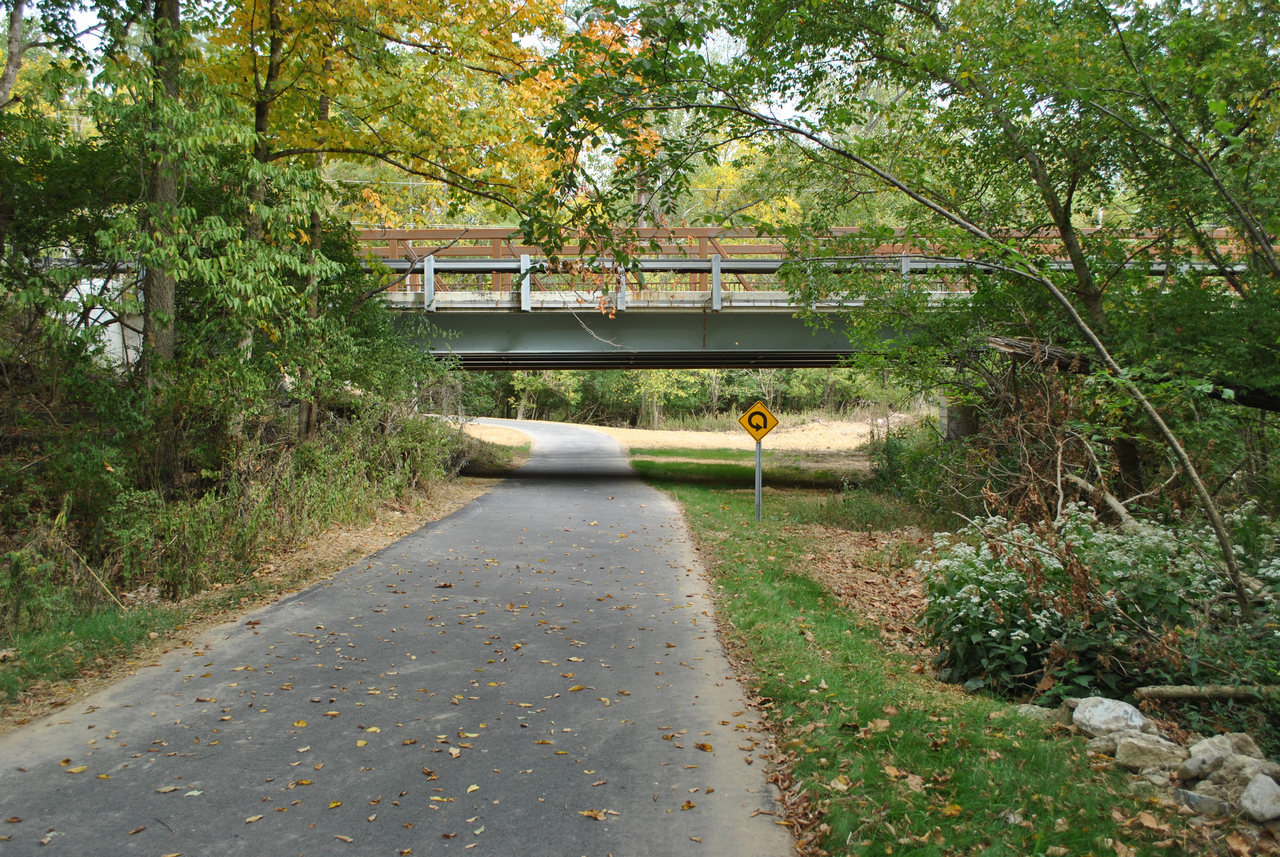 Path under a small bridge, lined with trees showing autumn colors.
