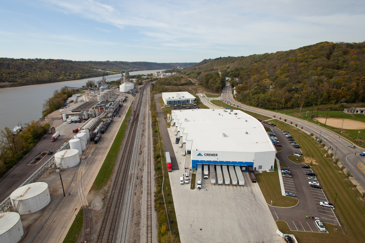 Aerial view of a white industrial building by a railway and river, surrounded by greenery.