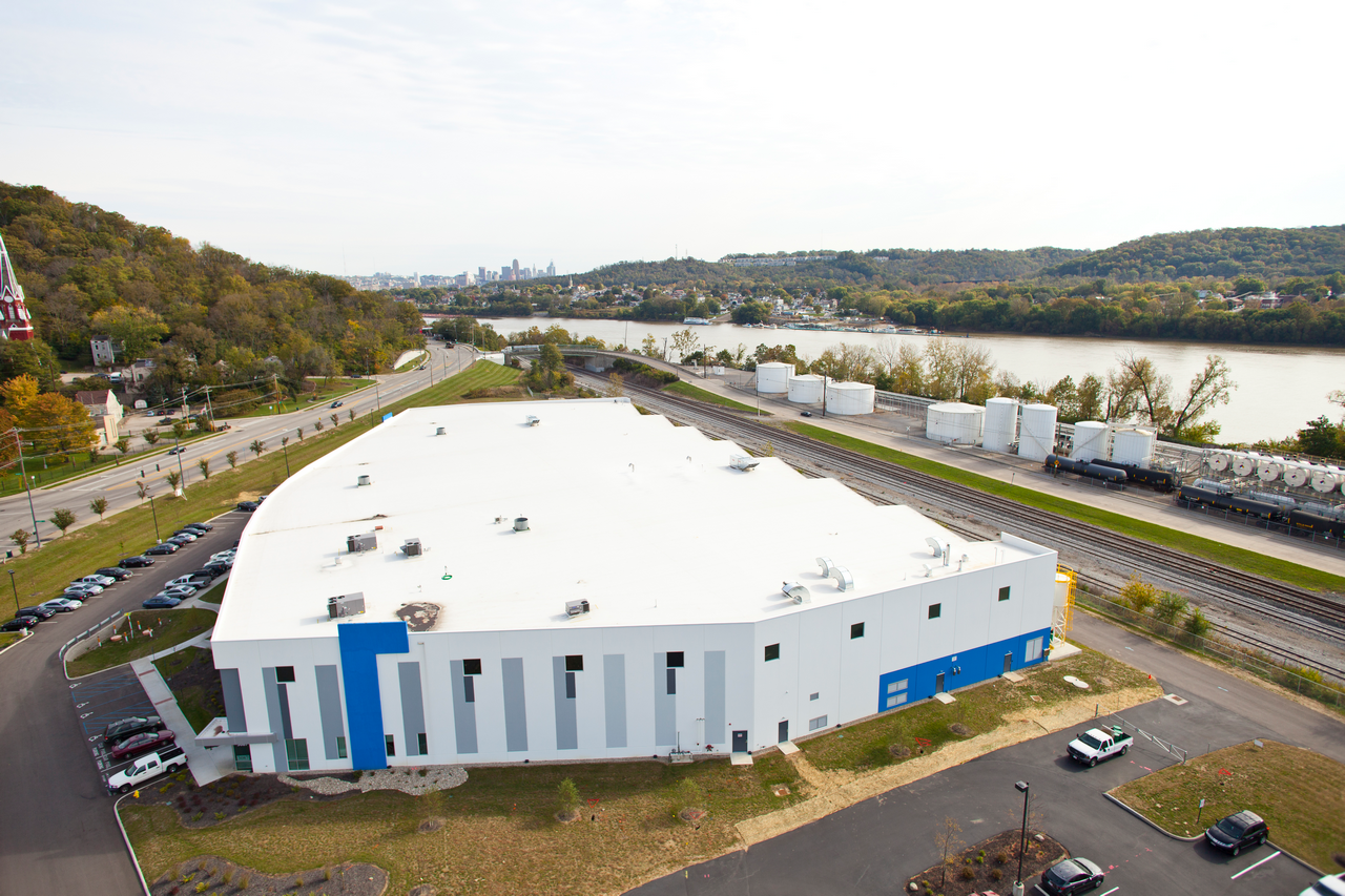 White industrial building near a river, surrounded by trees and train tracks.