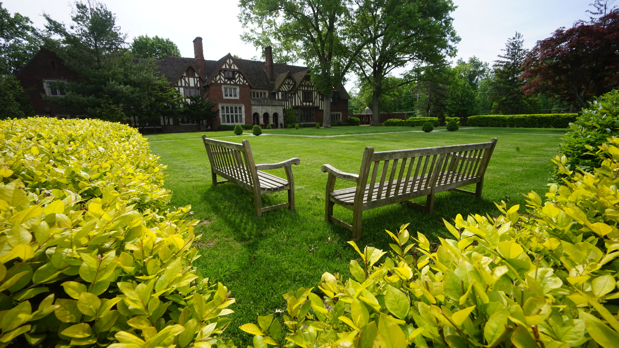 Benches on a manicured lawn, surrounded by lush greenery, with a large house in the background.