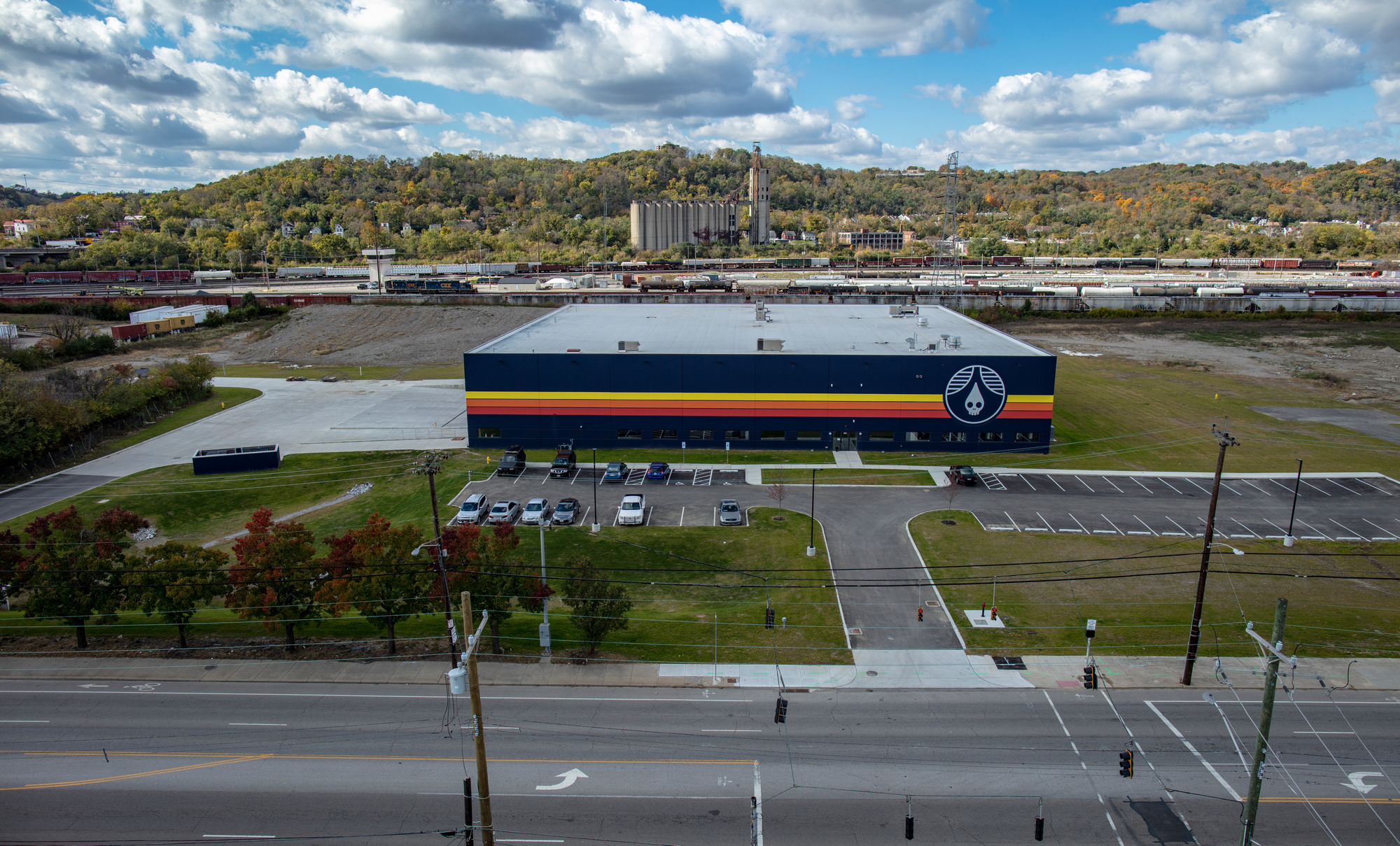 Warehouse with colorful stripes, parking lot in front, cloudy sky, trees, and hills in background.