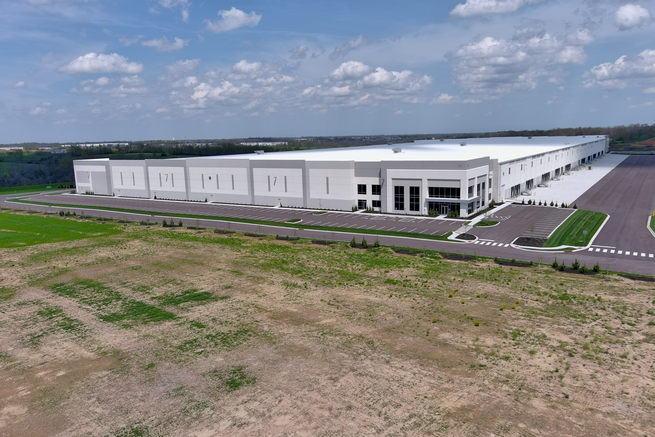 Large white warehouse with expansive parking lot under a partly cloudy sky.