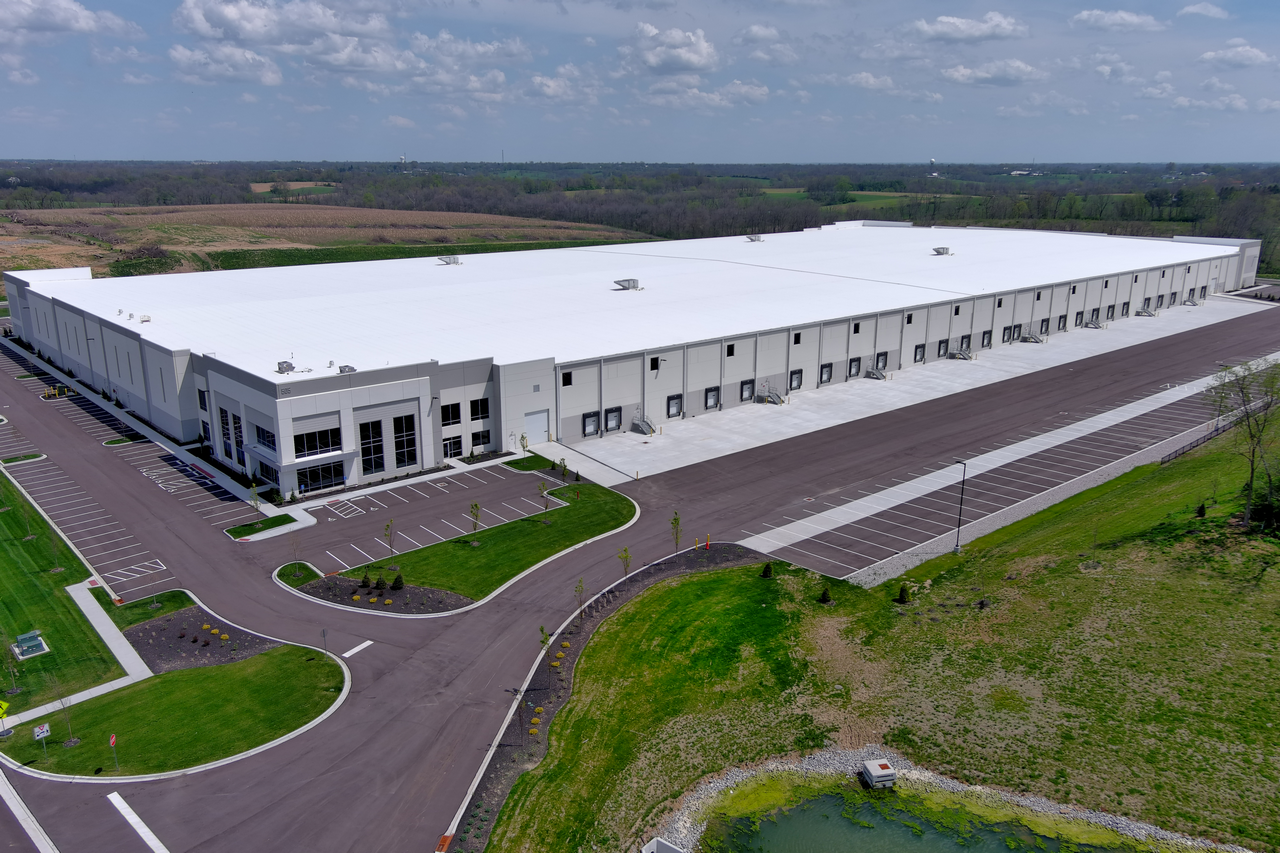 Large white warehouse with parking lot, surrounded by grass and fields under blue sky.
