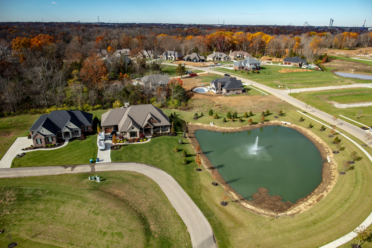 Aerial view of suburban homes by a pond with fountain, surrounded by autumn trees.