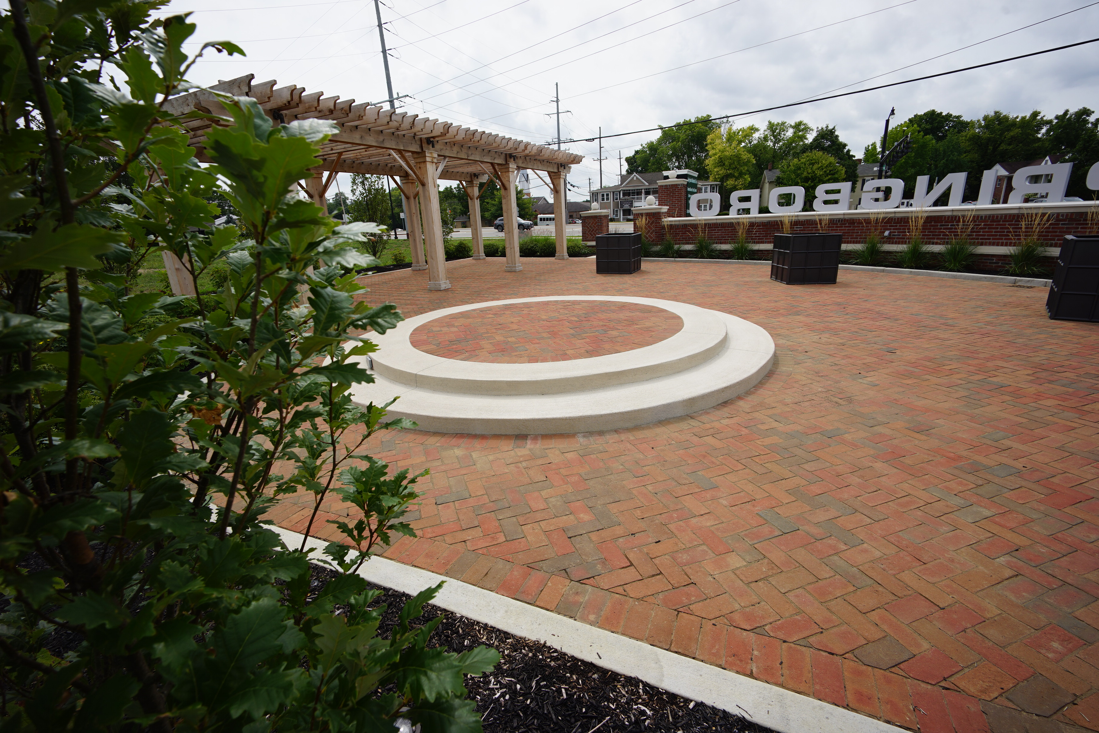 Brick plaza with a circular platform, pergola, and greenery.