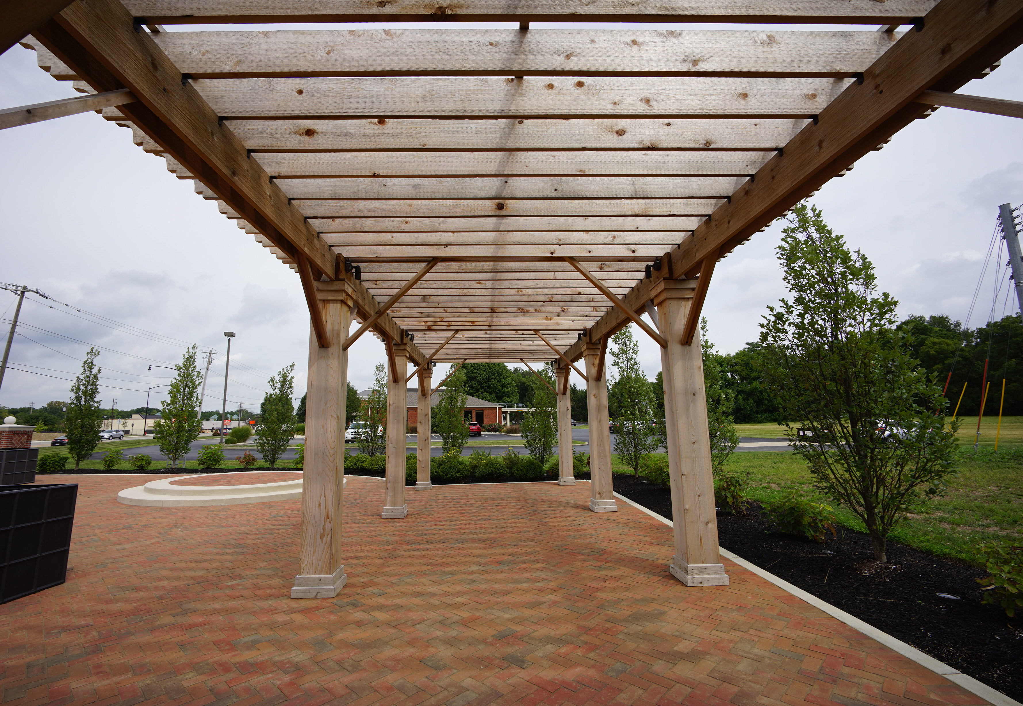 Wooden pergola over brick walkway with trees in the background.
