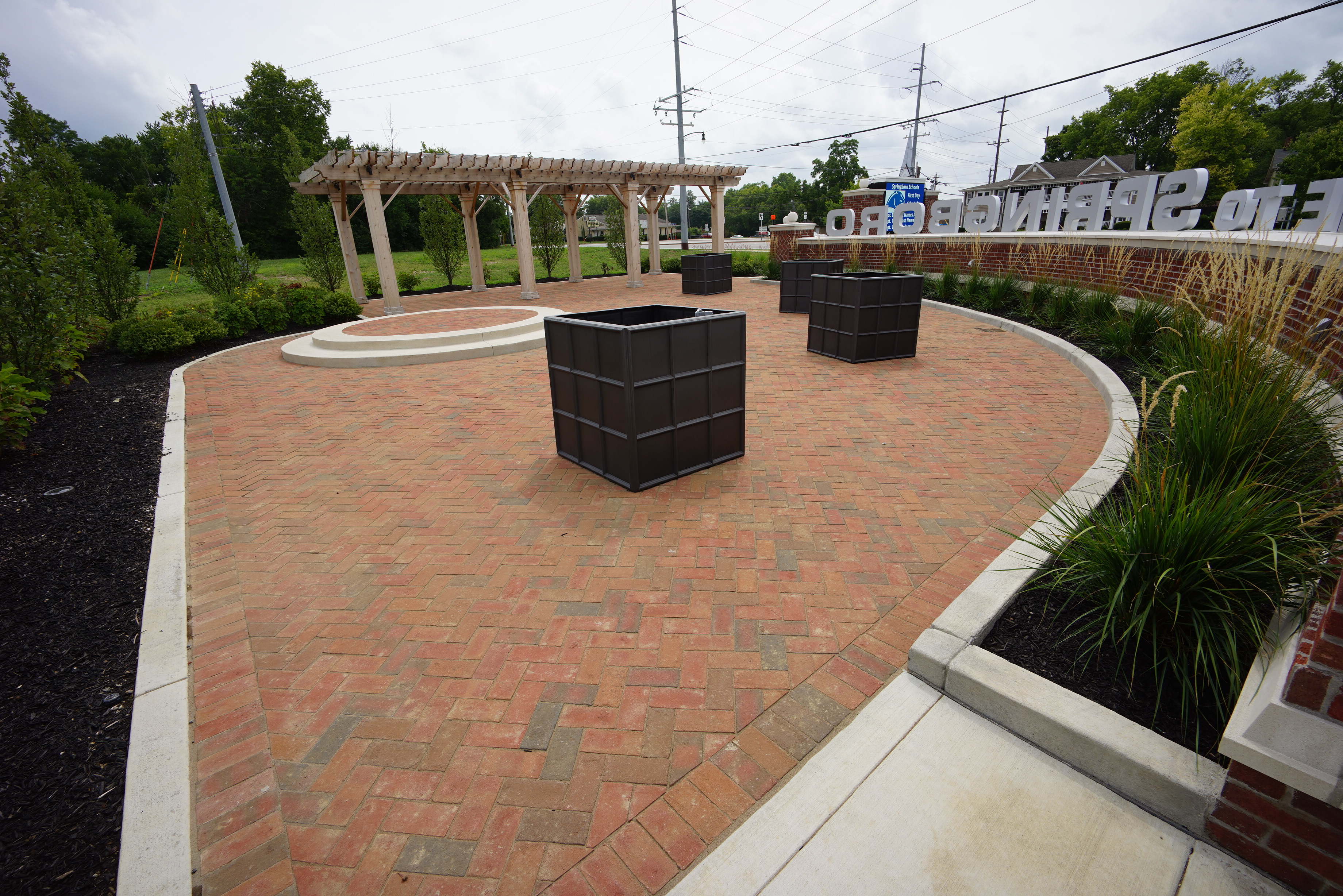 Brick pathway with pergola and planters, surrounded by greenery and a sign.