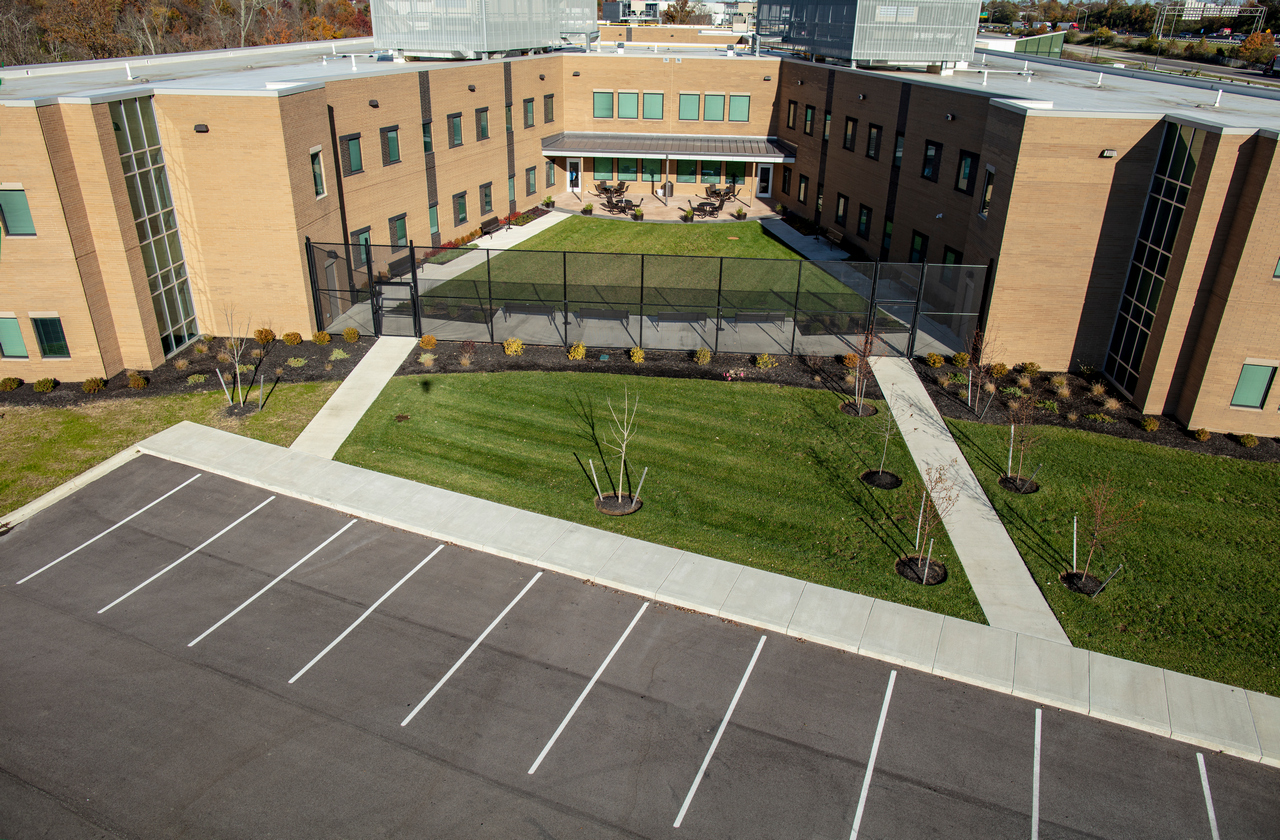 Brick building with courtyard, paths, and empty parking lot in front.