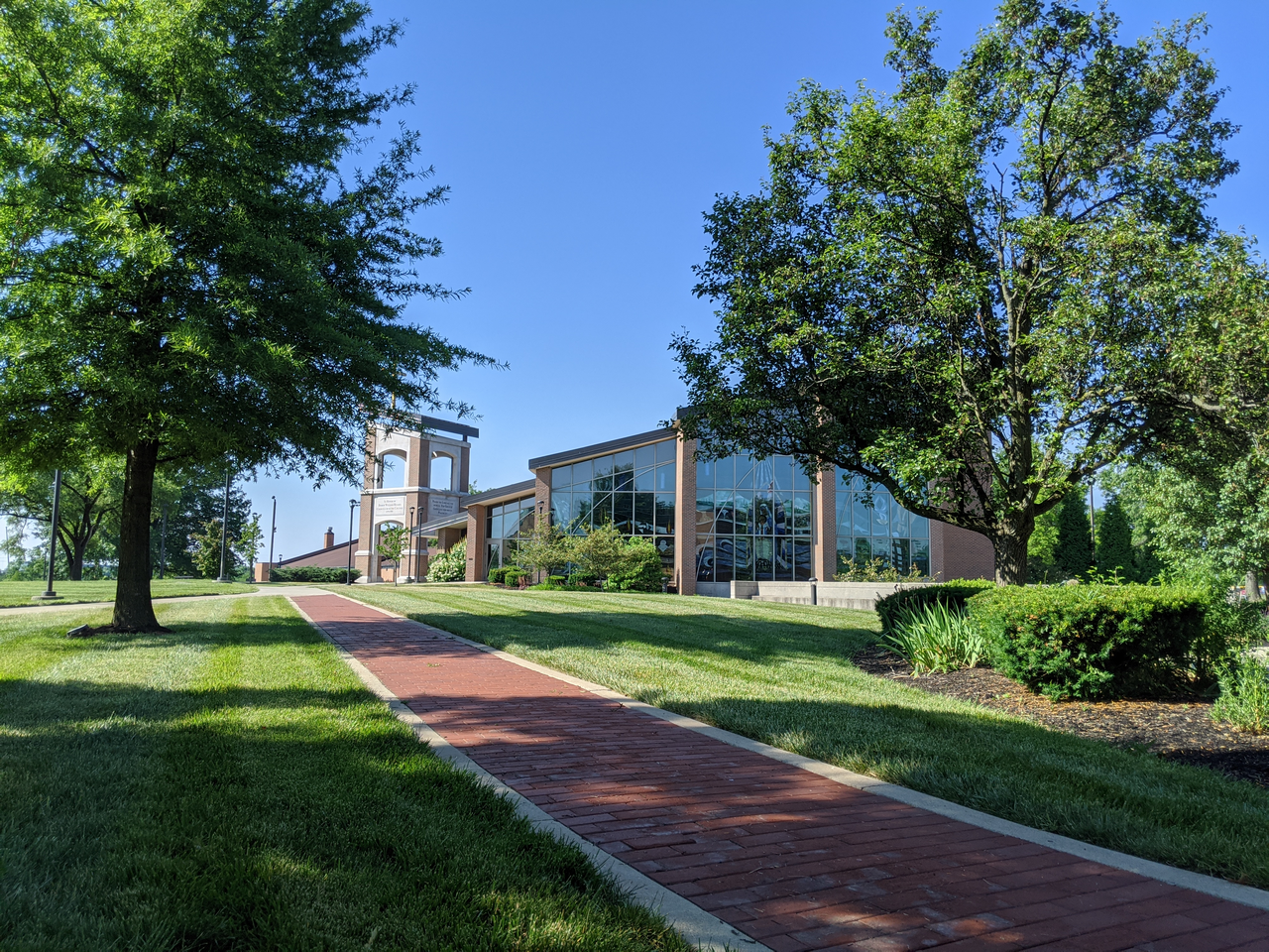 Brick path leading to a modern building, surrounded by green trees and grass.