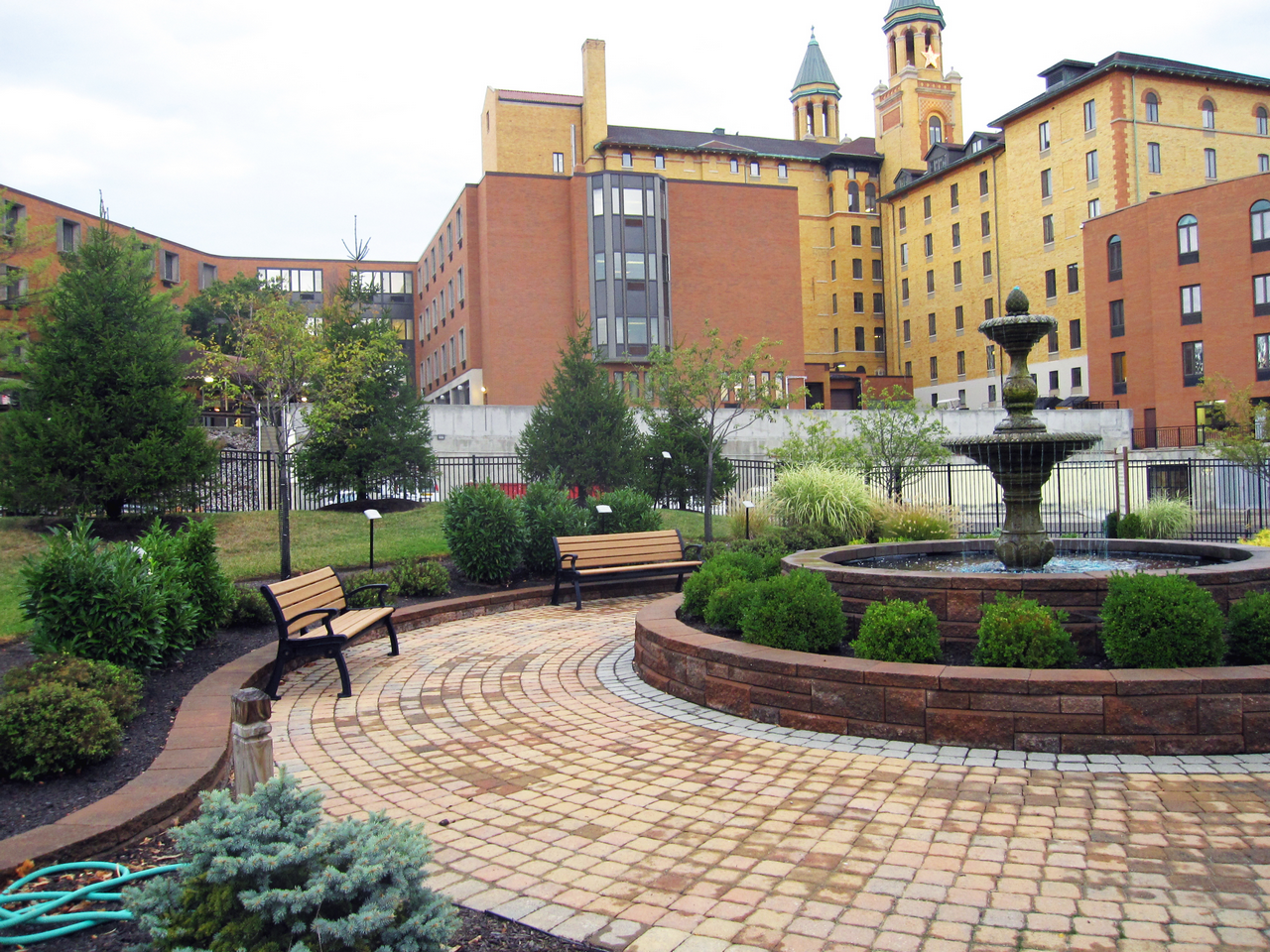 Brick-paved garden with benches and a fountain, surrounded by buildings.