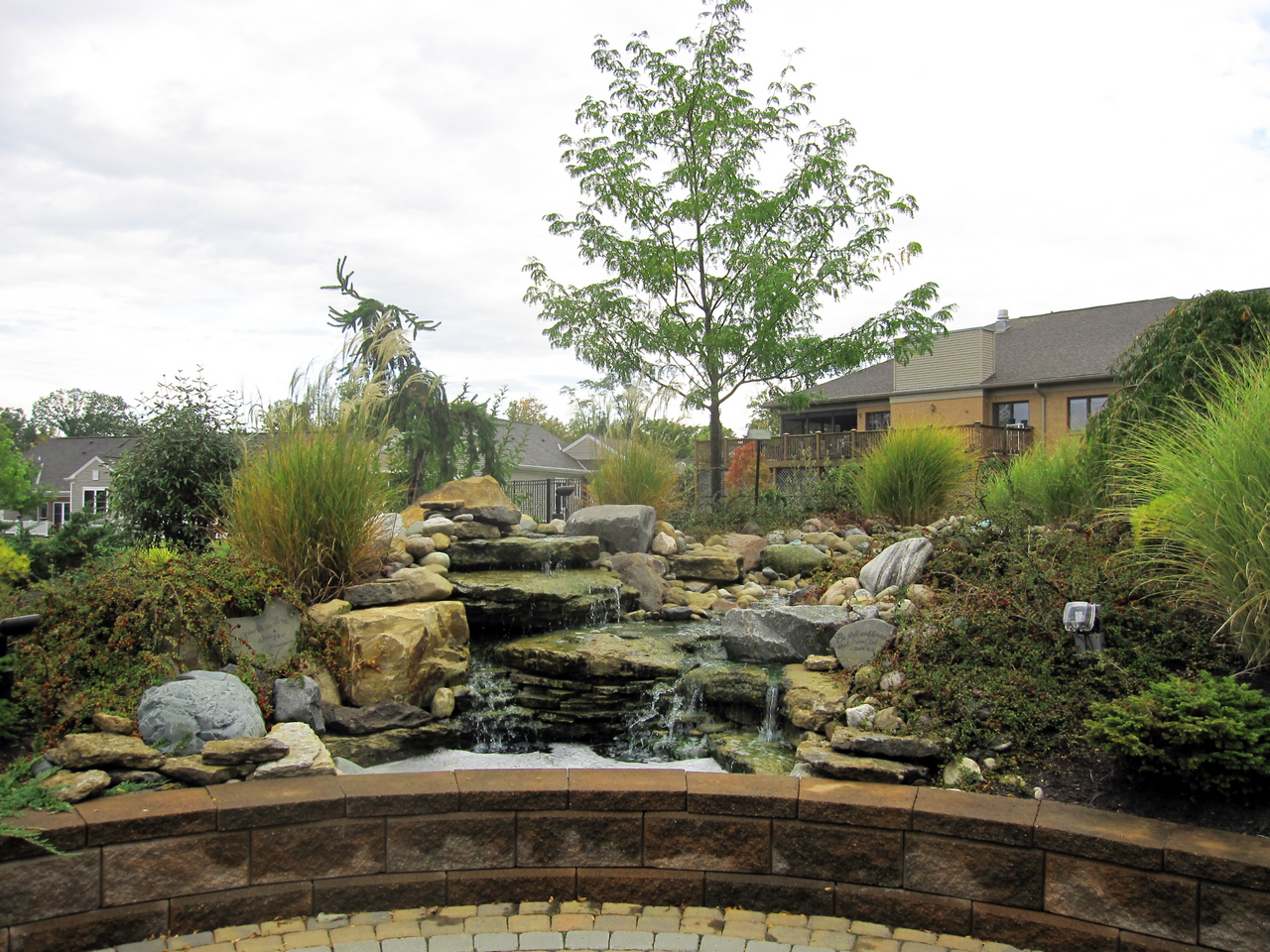 Garden with a stone pond, trees, and shrubs under a cloudy sky.