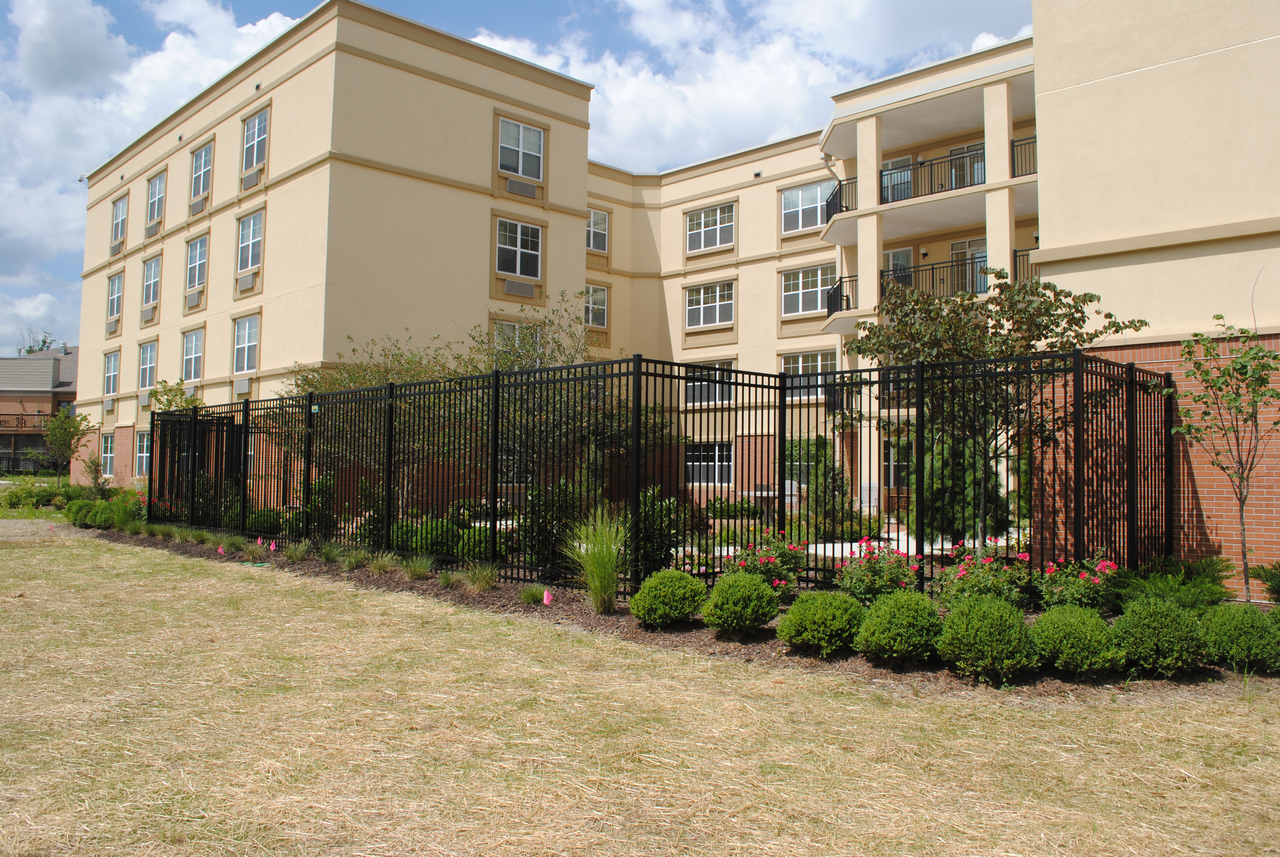 Four-story beige building with landscaped garden and black fence.