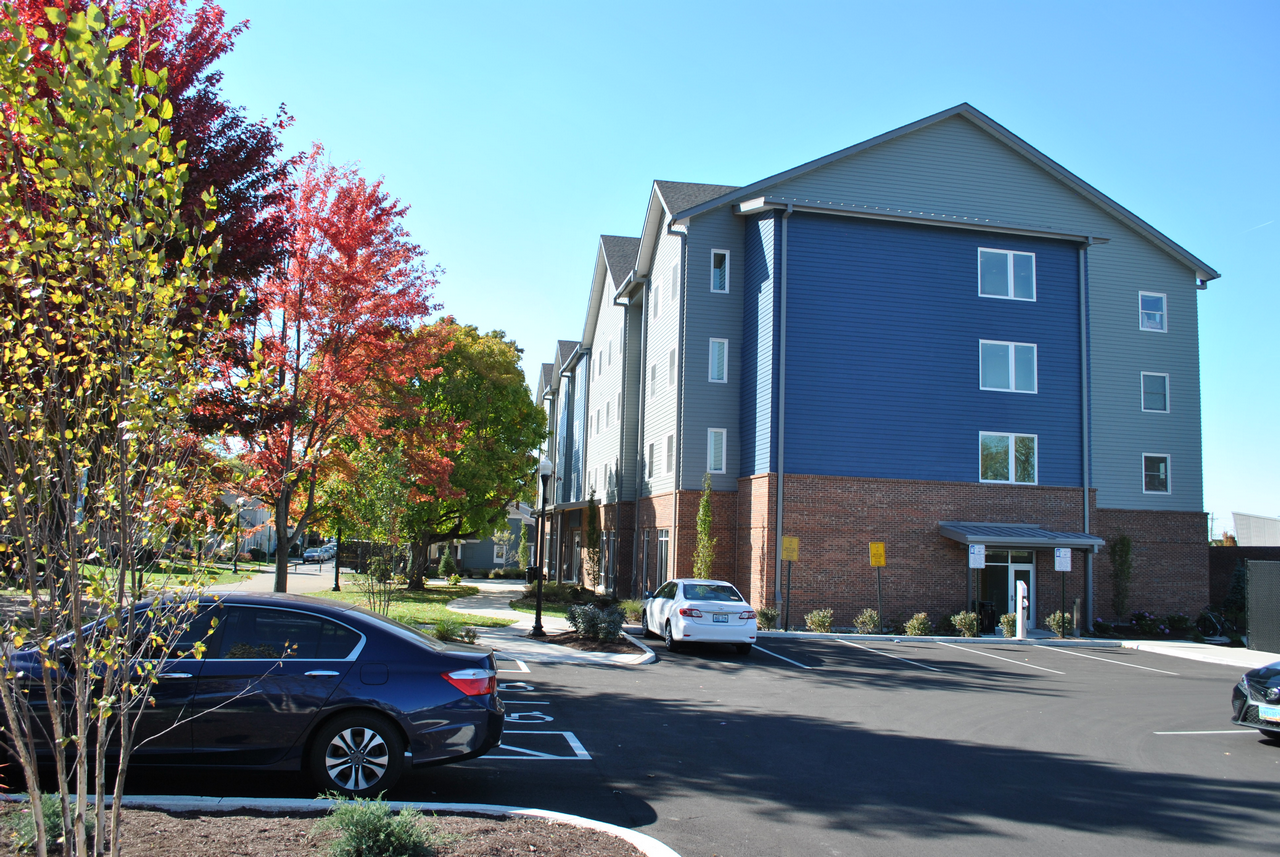 A blue and brick apartment building with parked cars and colorful autumn trees nearby.