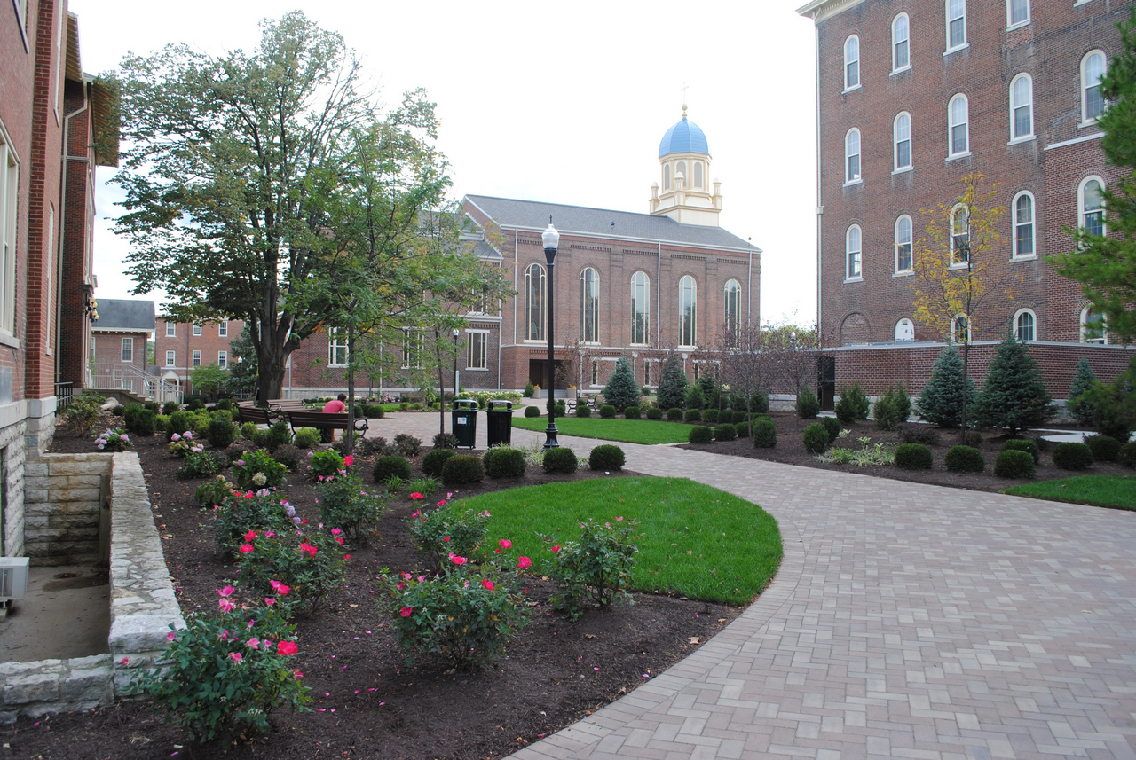Brick walkway through a garden with pink flowers, trees, and a building with a blue dome.