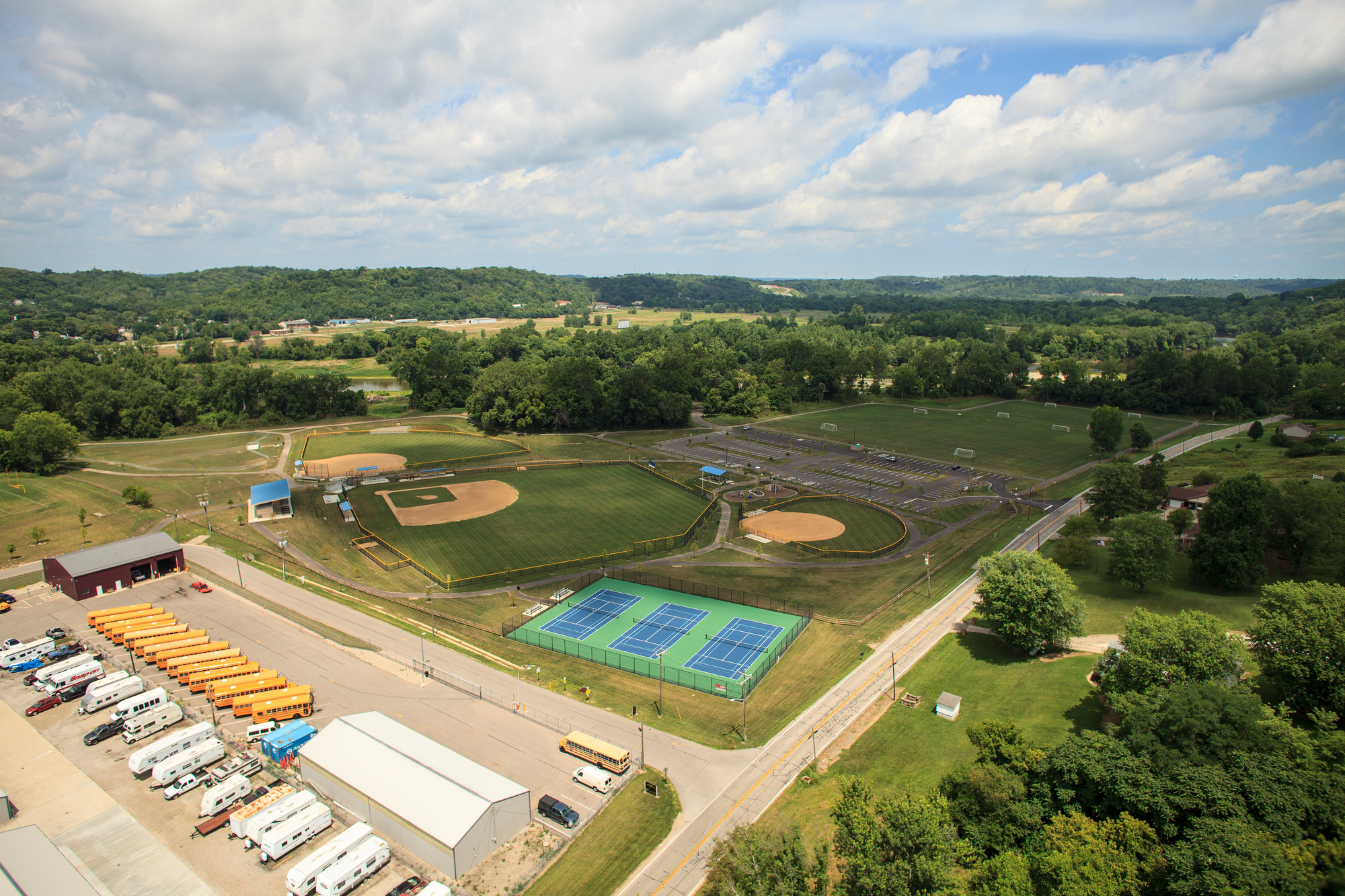 Aerial view of baseball fields, tennis courts, and surrounding greenery under a partly cloudy sky.