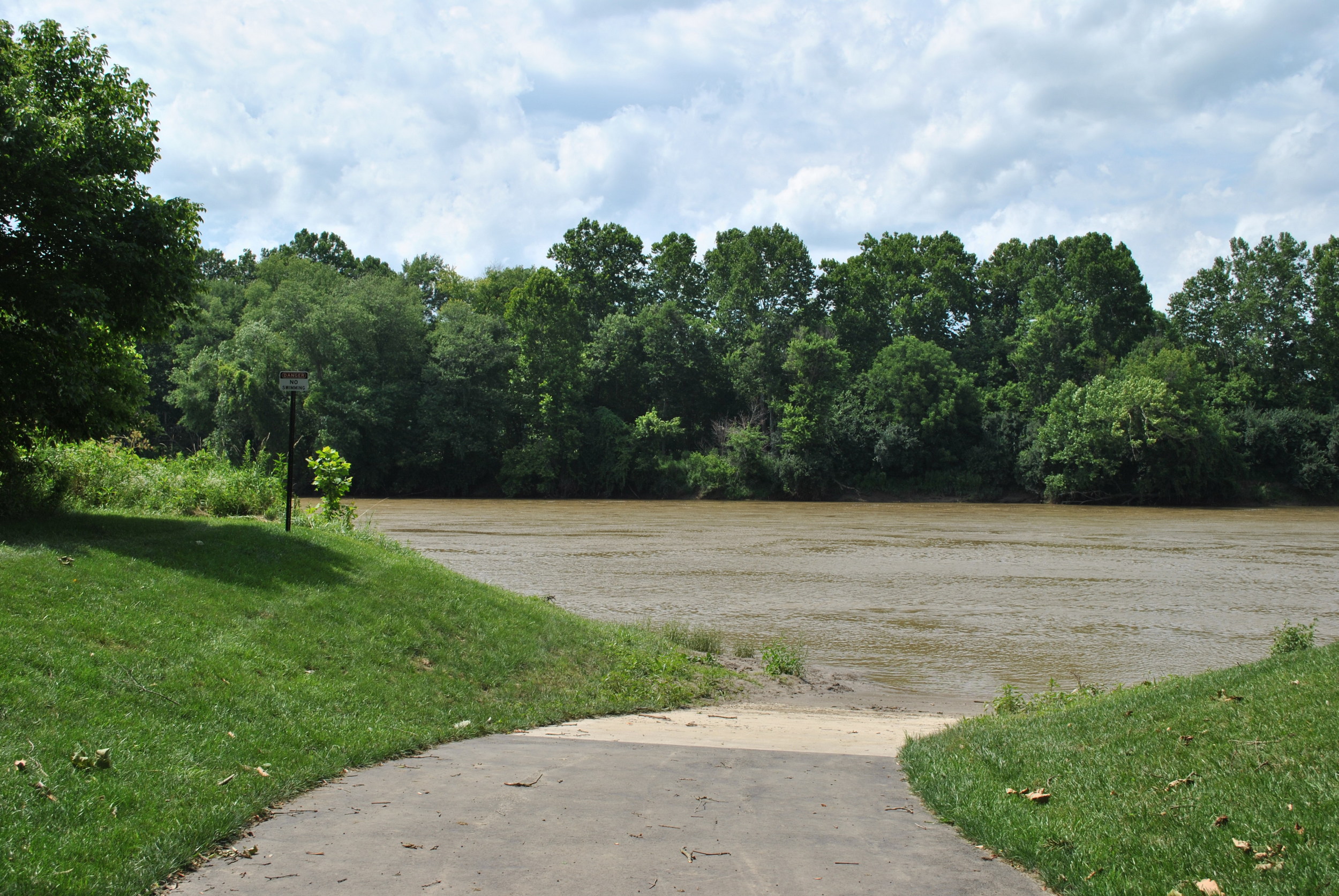 Path leading to a riverbank, surrounded by green trees under a cloudy sky.