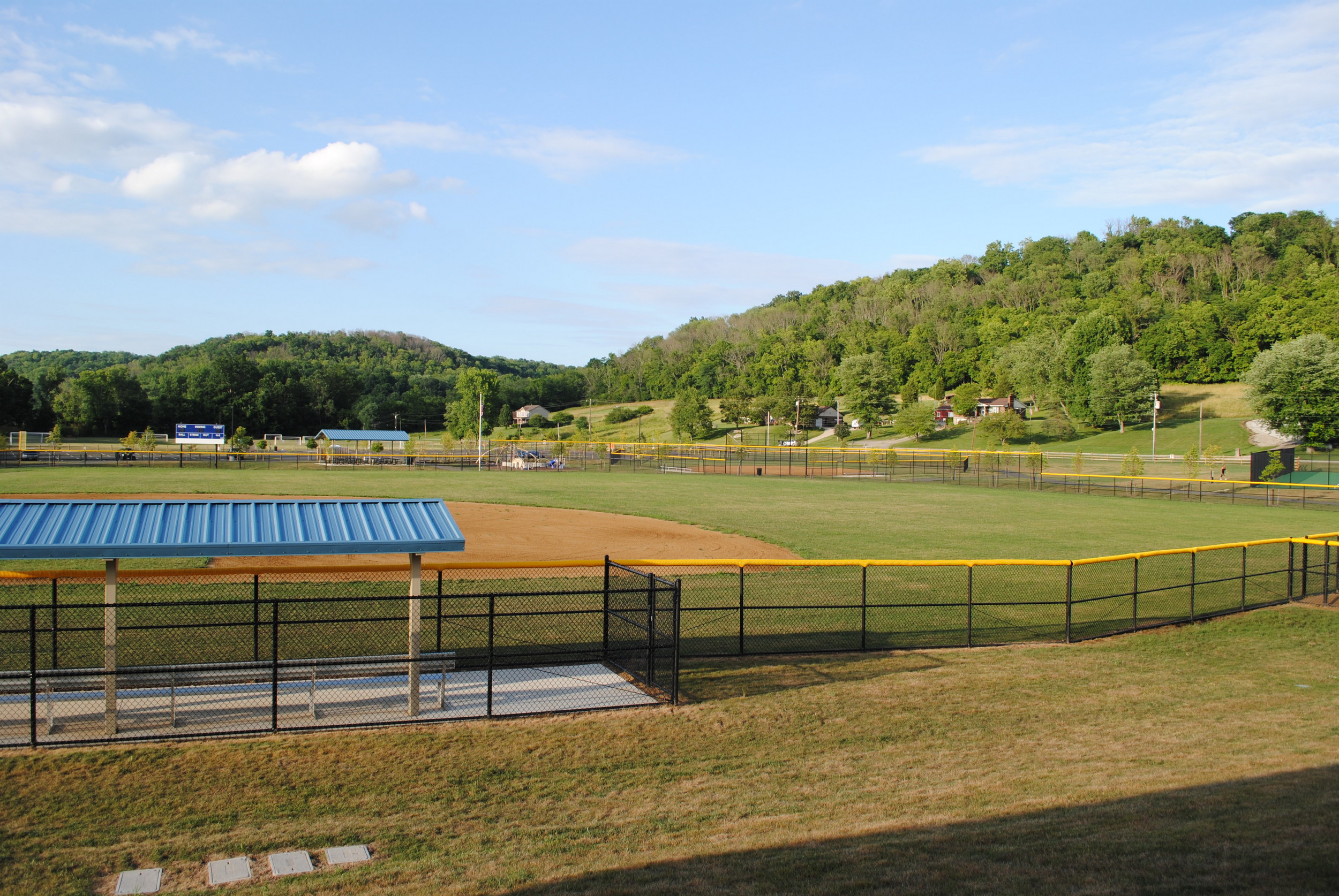 Sunny baseball field with blue dugouts, surrounded by trees and hills.