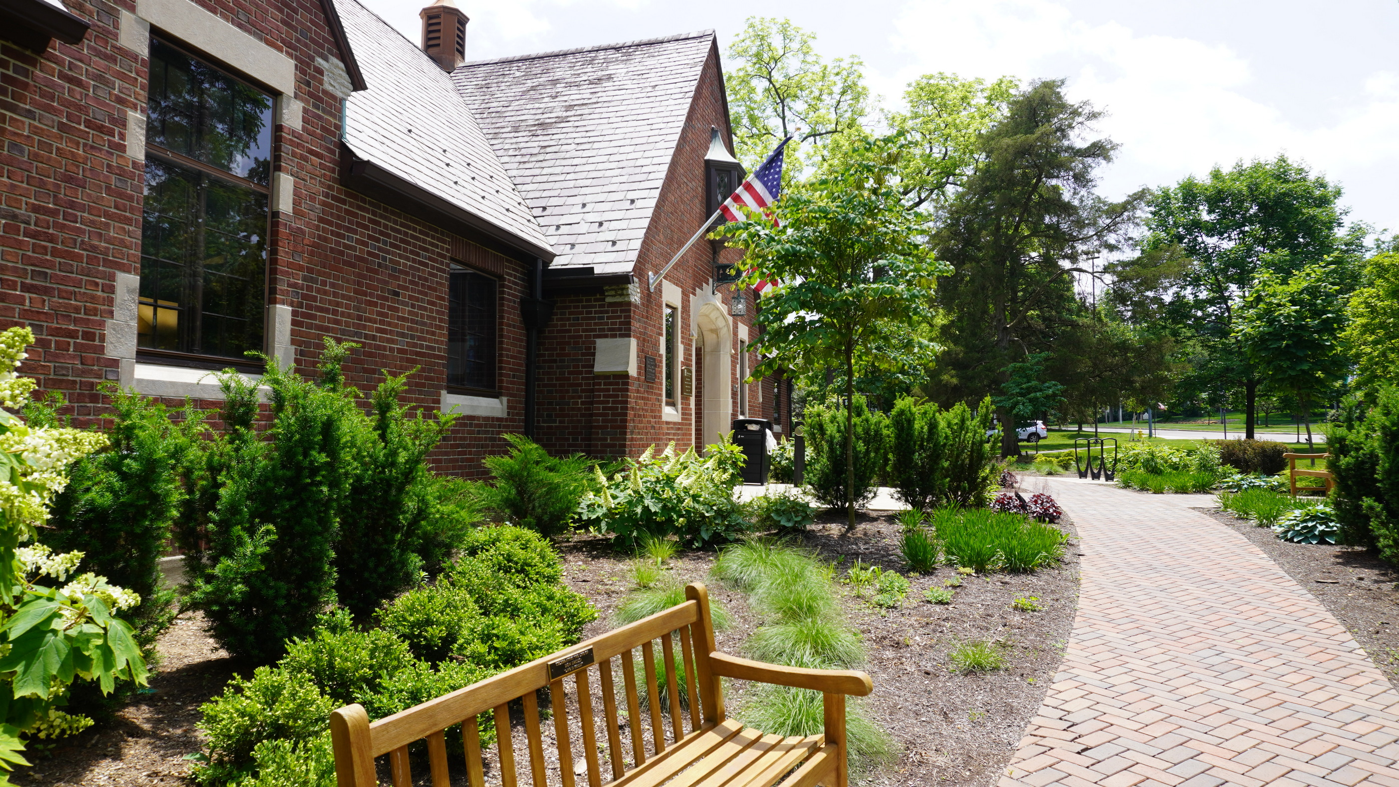 Brick path and bench beside a brick building with greenery and flags.