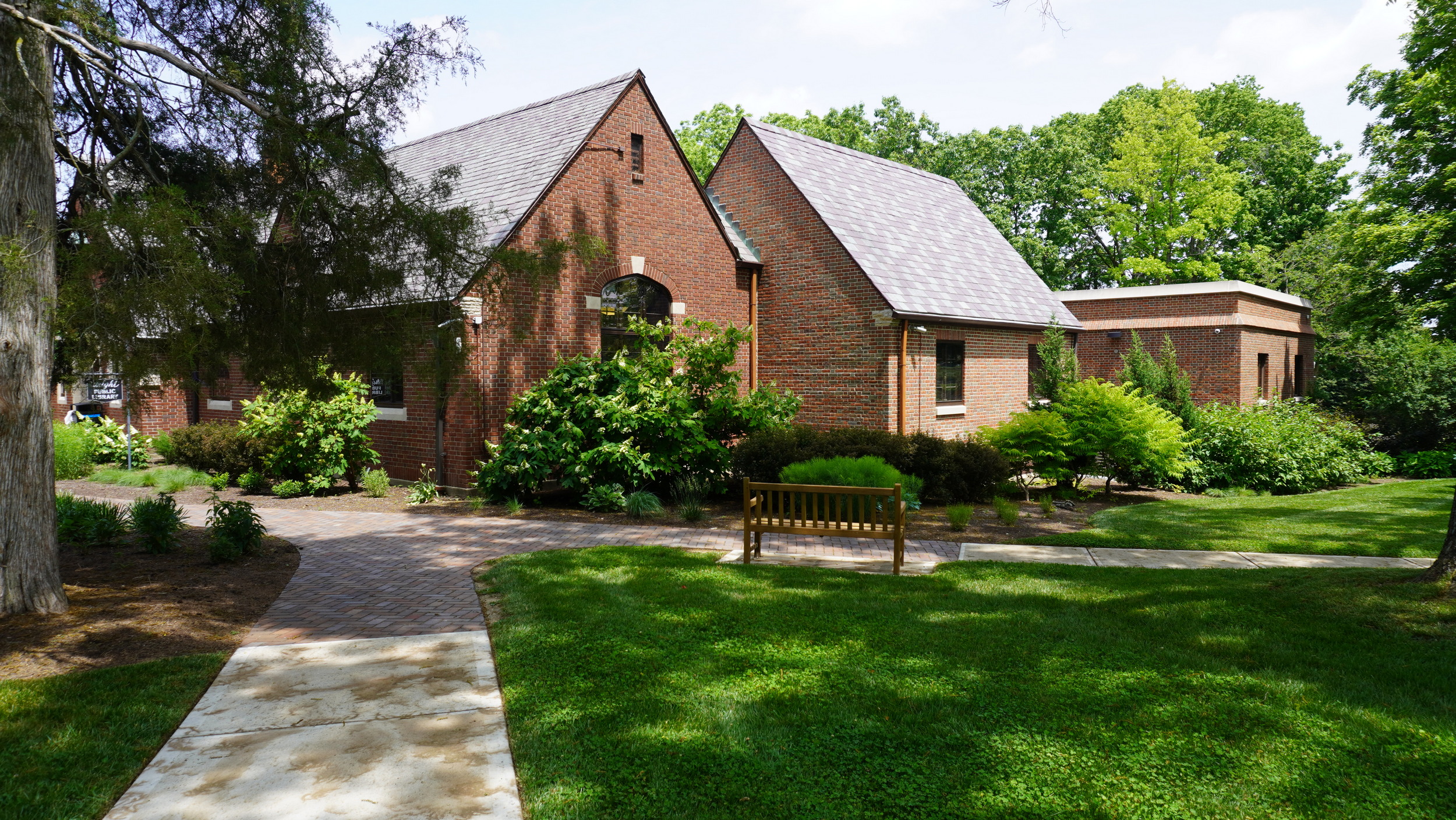 Brick building with gabled roofs, lush greenery, and a wooden bench in a garden setting.