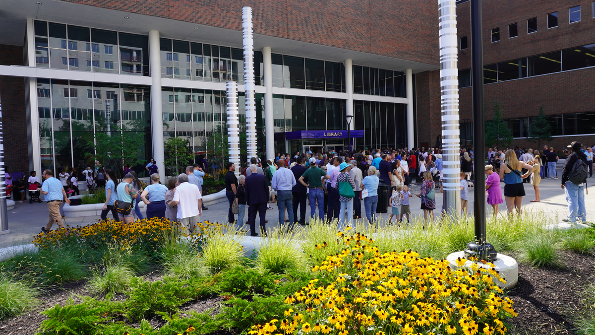 Crowd gathers outside a building with tall windows, surrounded by yellow flowers.