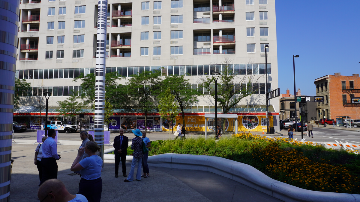 Street scene with people gathered near a flowerbed, high-rise building in the background.