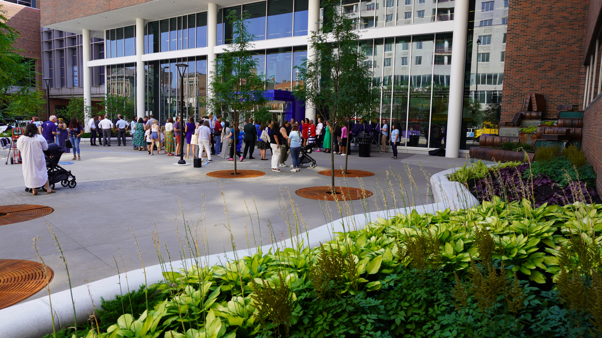 Crowd gathered outside modern building with garden and trees in foreground.