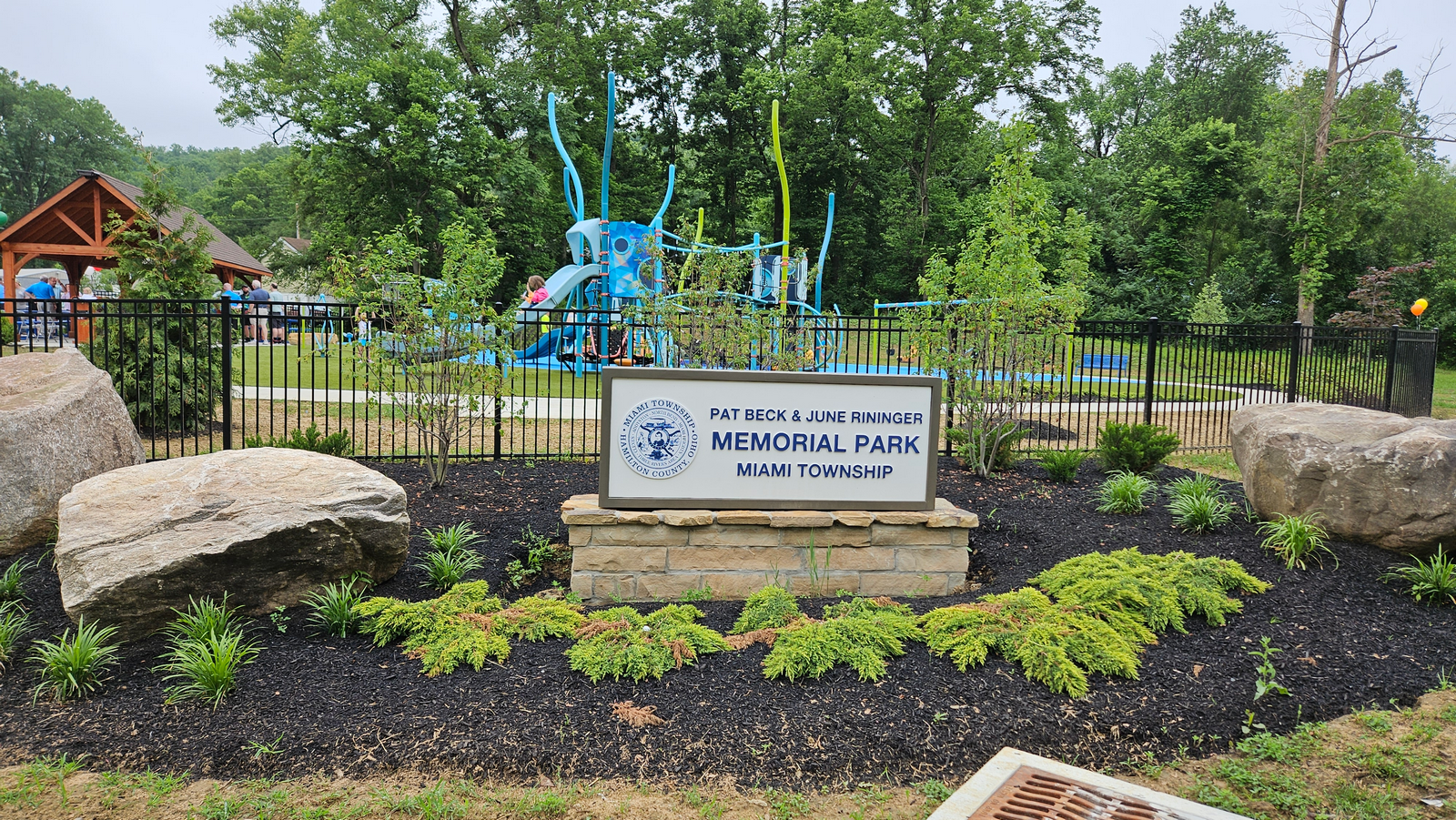 Park sign with rocks and greenery, playground equipment in the background.