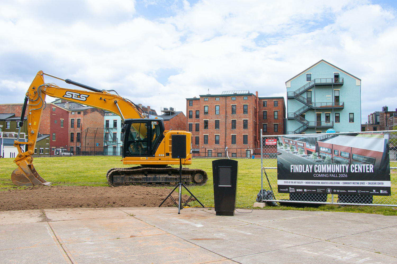 Findlay Community Center - Cincinnati, OH Yellow excavator on grass lot with city buildings in background.