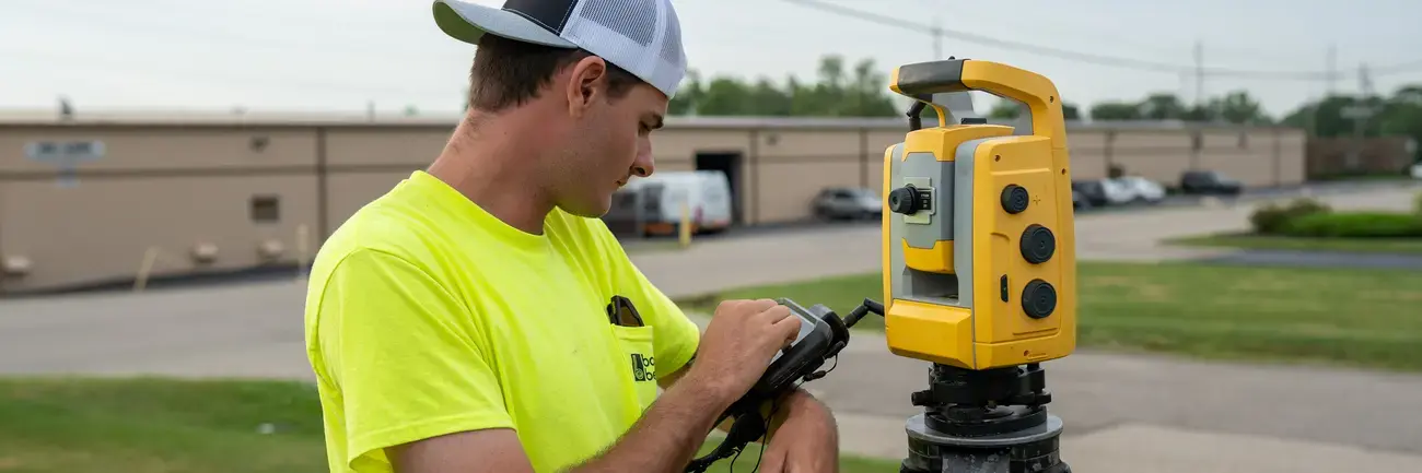 Man in yellow shirt adjusting survey equipment on tripod outdoors.